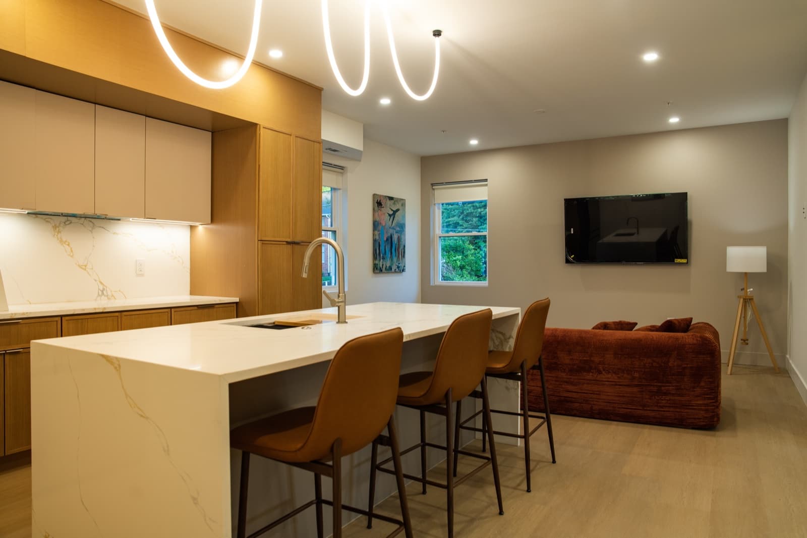 Open kitchen with marble waterfall island, four leather counter stools, signature squiggle pendant lighting, and the corduroy sectional beyond