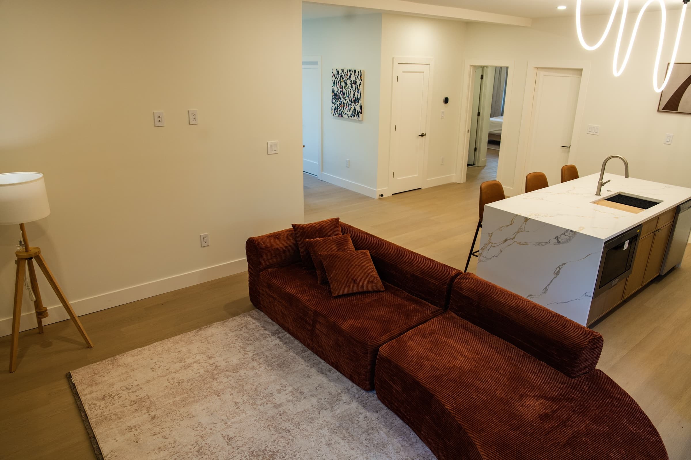 The Veyrai at Shadyside: corduroy sectional in the open living room with marble waterfall island, signature squiggle pendant lighting, and hallway art beyond